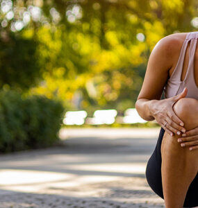 A young woman outdoors on a sunny day, leaning over and holding her knee in pain.