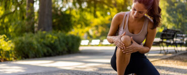 A young woman outdoors on a sunny day, leaning over and holding her knee in pain.