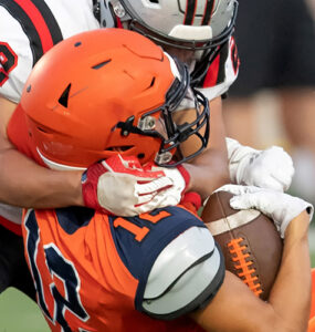 A football player wearing orange being tackled by a player wearing white, both about to hit the ground.