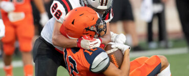 A football player wearing orange being tackled by a player wearing white, both about to hit the ground.