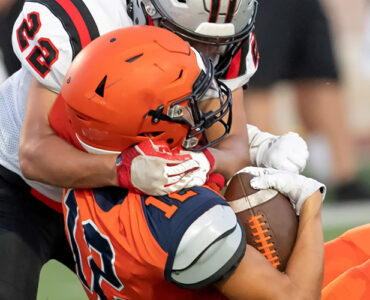A football player wearing orange being tackled by a player wearing white, both about to hit the ground.