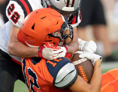 A football player wearing orange being tackled by a player wearing white, both about to hit the ground.