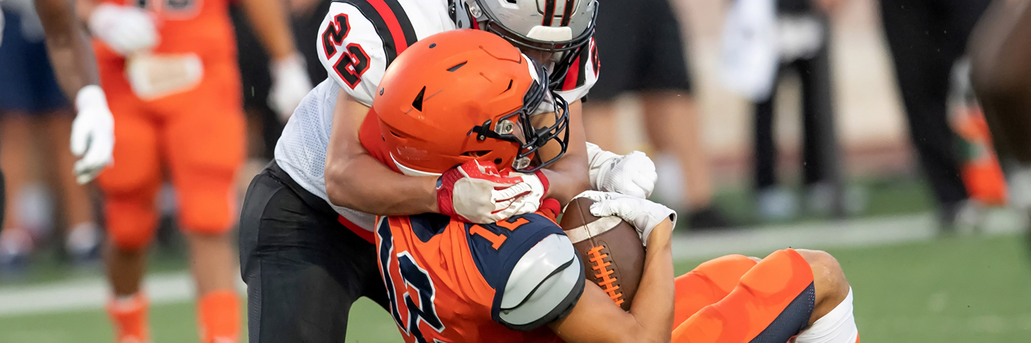 A football player wearing orange being tackled by a player wearing white, both about to hit the ground.