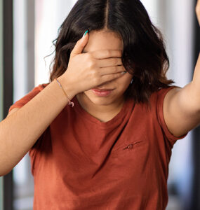 A young woman standing in her bathroom, covering her eyes because she's feeling dizzy.