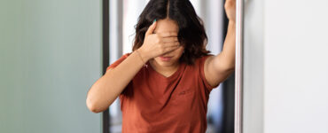 A young woman standing in her bathroom, covering her eyes because she's feeling dizzy.