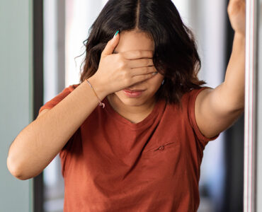 A young woman standing in her bathroom, covering her eyes because she's feeling dizzy.
