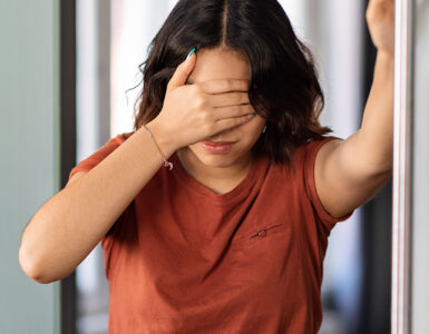 A young woman standing in her bathroom, covering her eyes because she's feeling dizzy.