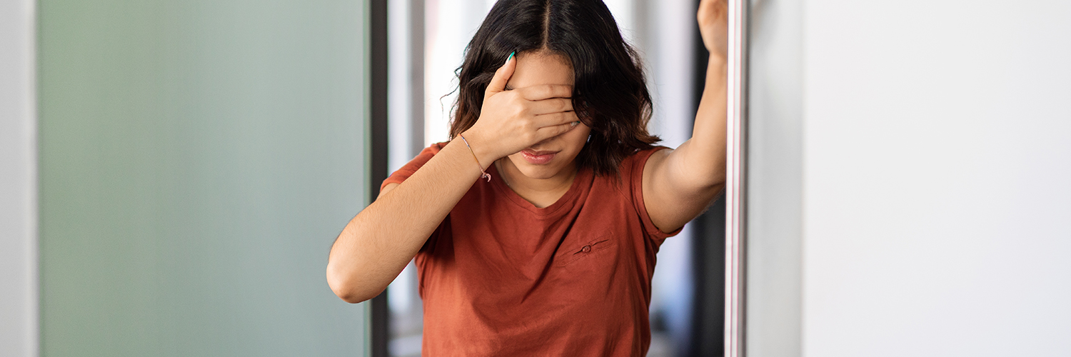 A young woman standing in her bathroom, covering her eyes because she's feeling dizzy.