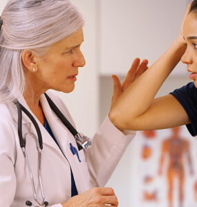A teenage girl in a sports uniform with a female doctor in her office, holding her head in pain.