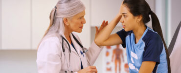 A teenage girl in a sports uniform with a female doctor in her office, holding her head in pain.
