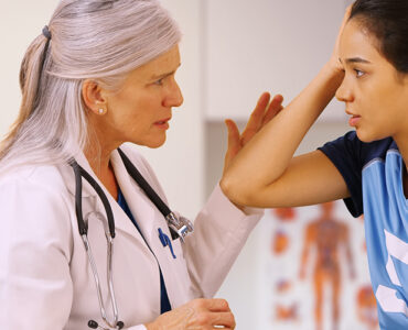 A teenage girl in a sports uniform with a female doctor in her office, holding her head in pain.