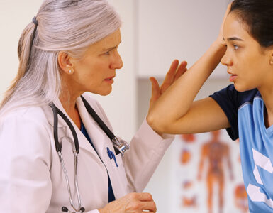 A teenage girl in a sports uniform with a female doctor in her office, holding her head in pain.