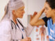 A teenage girl in a sports uniform with a female doctor in her office, holding her head in pain.