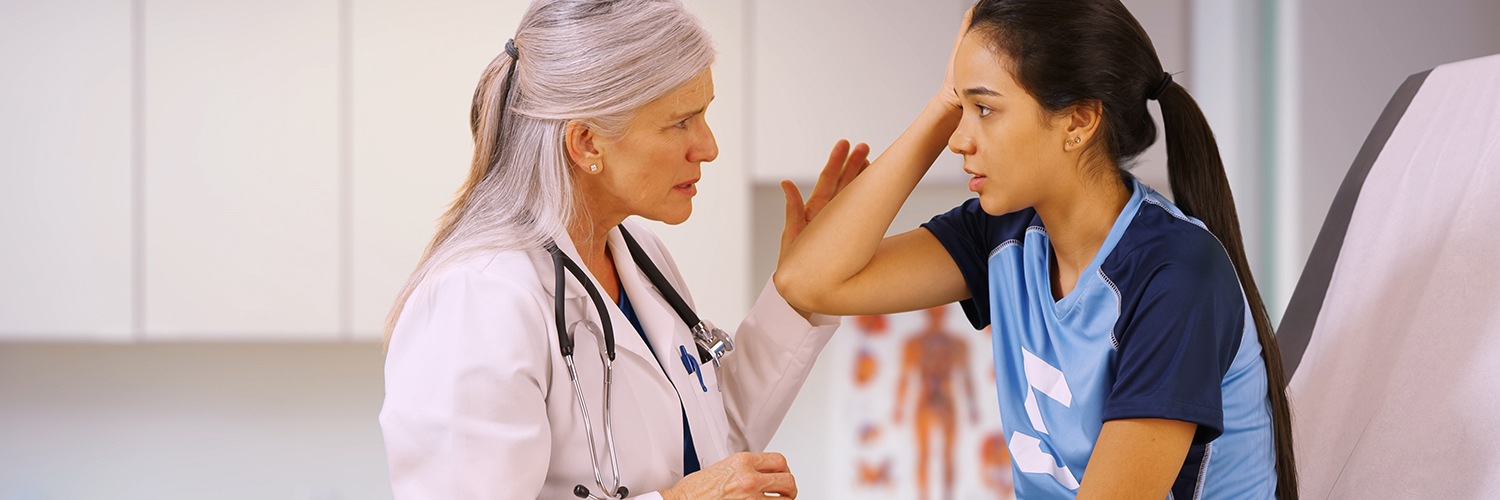 A teenage girl in a sports uniform with a female doctor in her office, holding her head in pain.
