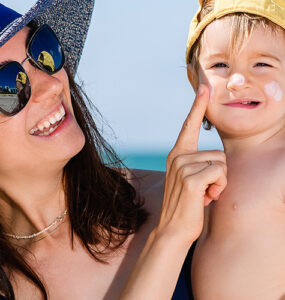 A mother wearing a sun hat and sunglasses at the beach, holding her son and applying sunscreen to his cheeks.