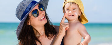 A mother wearing a sun hat and sunglasses at the beach, holding her son and applying sunscreen to his cheeks.