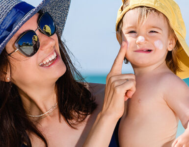 A mother wearing a sun hat and sunglasses at the beach, holding her son and applying sunscreen to his cheeks.