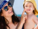 A mother wearing a sun hat and sunglasses at the beach, holding her son and applying sunscreen to his cheeks.