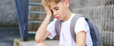 A young boy at school, sitting alone looking sad with his hand on his head.