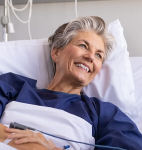 An older female patient sitting up in a hospital bed, smiling at a young man next to her.