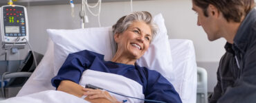 An older female patient sitting up in a hospital bed, smiling at a young man next to her.