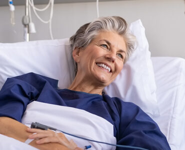 An older female patient sitting up in a hospital bed, smiling at a young man next to her.