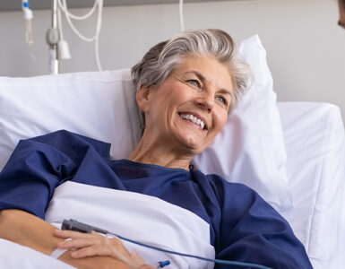 An older female patient sitting up in a hospital bed, smiling at a young man next to her.