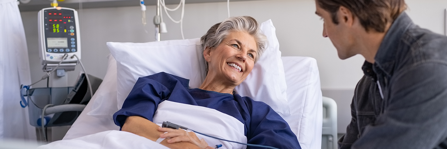 An older female patient sitting up in a hospital bed, smiling at a young man next to her.
