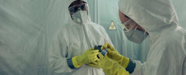 Two doctors standing together, wearing white protective clothing, with masks, gloves and goggles.
