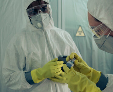 Two doctors standing together, wearing white protective clothing, with masks, gloves and goggles.
