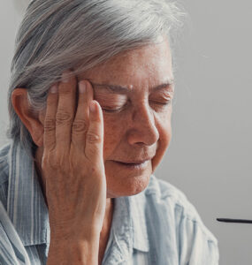 Elderly woman sits on a sofa at home suffering from a headache.