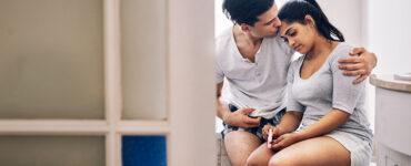 A woman sitting in a bathroom holding a pregnancy test and looking sad while a man kisses her forehead.