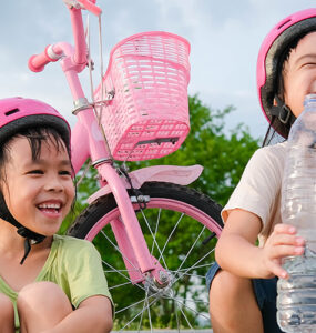 Little girls with helmets on taking a break from riding scooter and bike, drinking a water