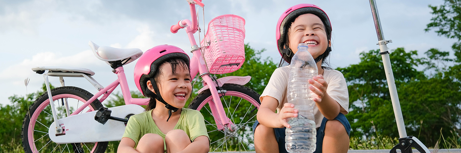 Little girls with helmets on taking a break from riding scooter and bike, drinking a water
