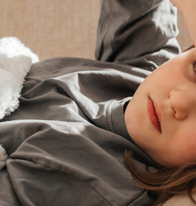 A young girl laying on a couch, holding her forehead while looking sickly.
