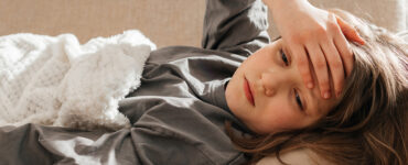 A young girl laying on a couch, holding her forehead while looking sickly.