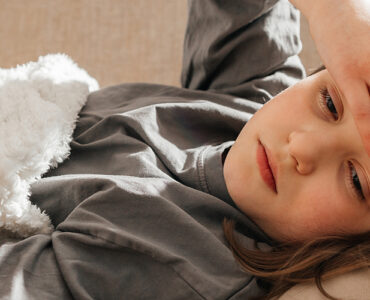 A young girl laying on a couch, holding her forehead while looking sickly.
