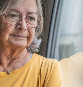 Elderly woman in a yellow shirt stands by a window with a reflective gaze.