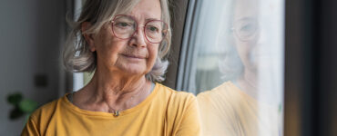 Elderly woman in a yellow shirt stands by a window with a reflective gaze.