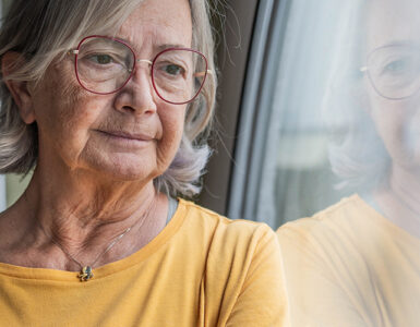 Elderly woman in a yellow shirt stands by a window with a reflective gaze.