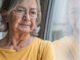 Elderly woman in a yellow shirt stands by a window with a reflective gaze.