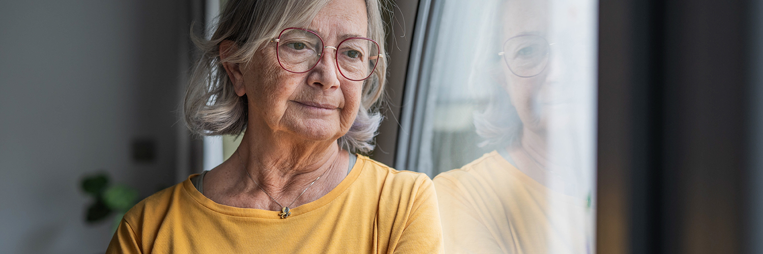 Elderly woman in a yellow shirt stands by a window with a reflective gaze.
