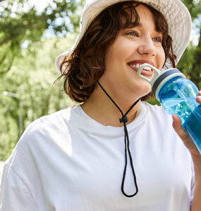 A fit young woman is exercising outdoors, happily sipping water and smiling while walking on a path.