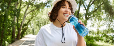 A fit young woman is exercising outdoors, happily sipping water and smiling while walking on a path.