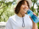 A fit young woman is exercising outdoors, happily sipping water and smiling while walking on a path.