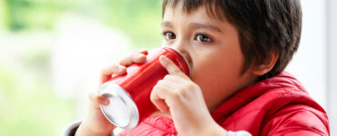 A young boy wearing a red coat and sipping from a red soda can.