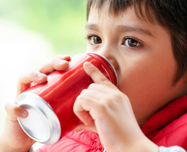 A young boy wearing a red coat and sipping from a red soda can.