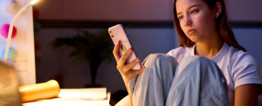 Teenage girl sitting at a desk in her bedroom at night looking at a mobile phone, illustrating teen screen time.