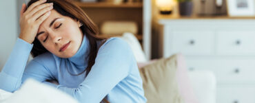 A woman in her 40s wearing a blue sweater and sitting on her couch, holding her forehead and looking tired.
