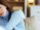 A woman in her 40s wearing a blue sweater and sitting on her couch, holding her forehead and looking tired.